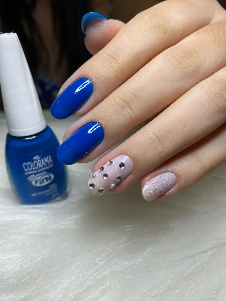 Unhas azul cobalto com duas unhas claras decoradas com pedrarias, ao lado do esmalte Colorama